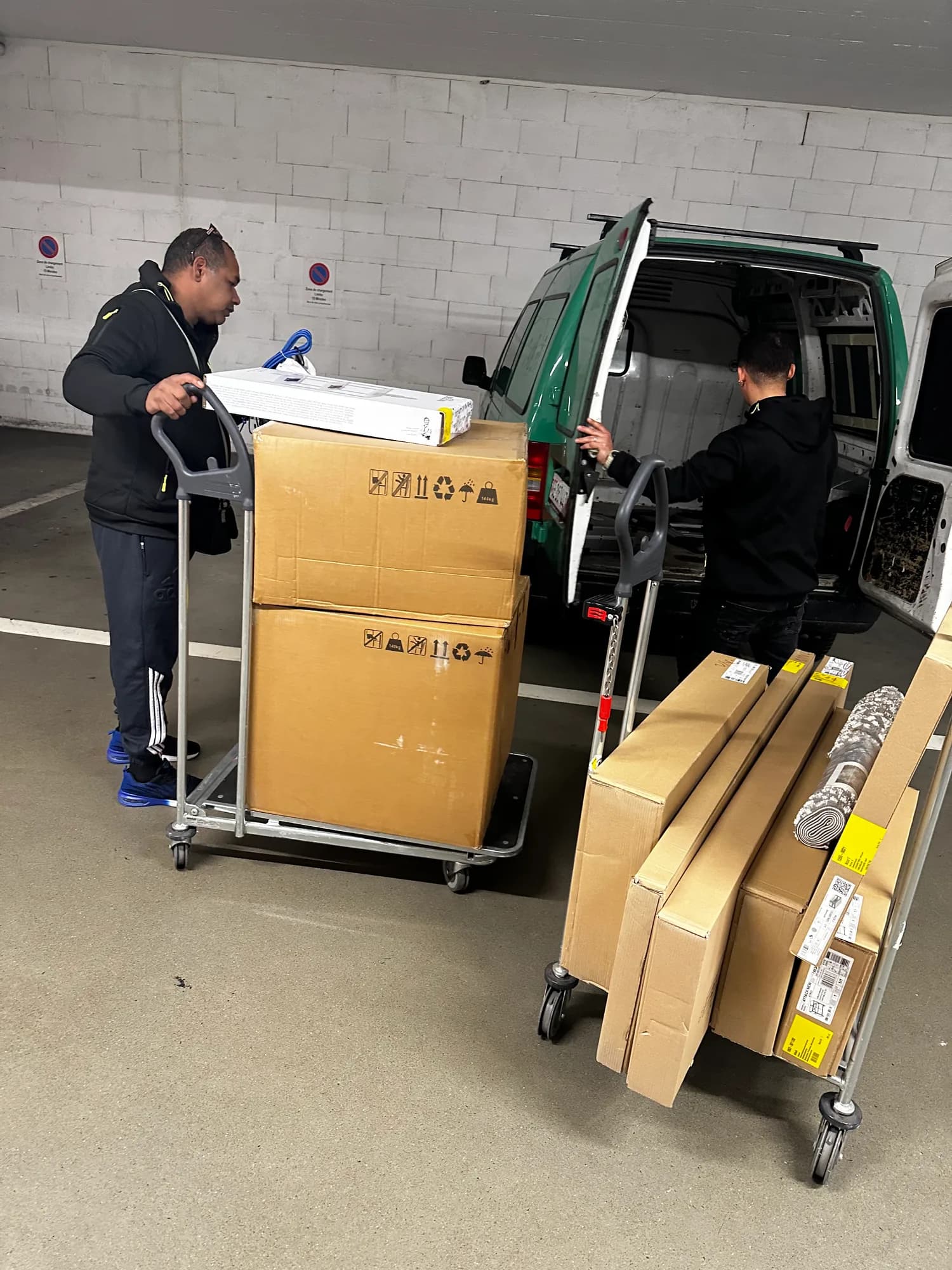 Deux ouvriers chargent des cartons sur des chariots devant un utilitaire, dans un parking