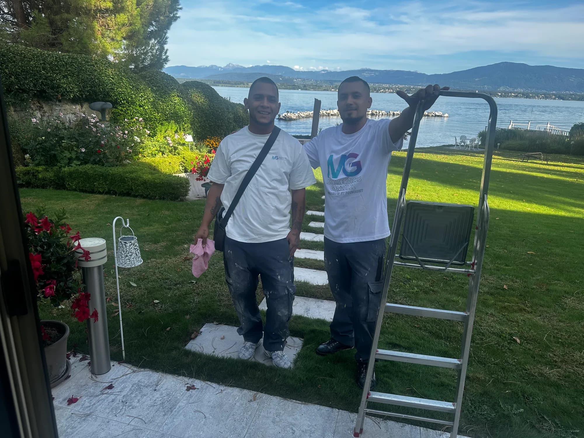 Deux ouvriers de l'équipe en t-shirt MG, une échelle à la main, dans un jardin au bord du lac