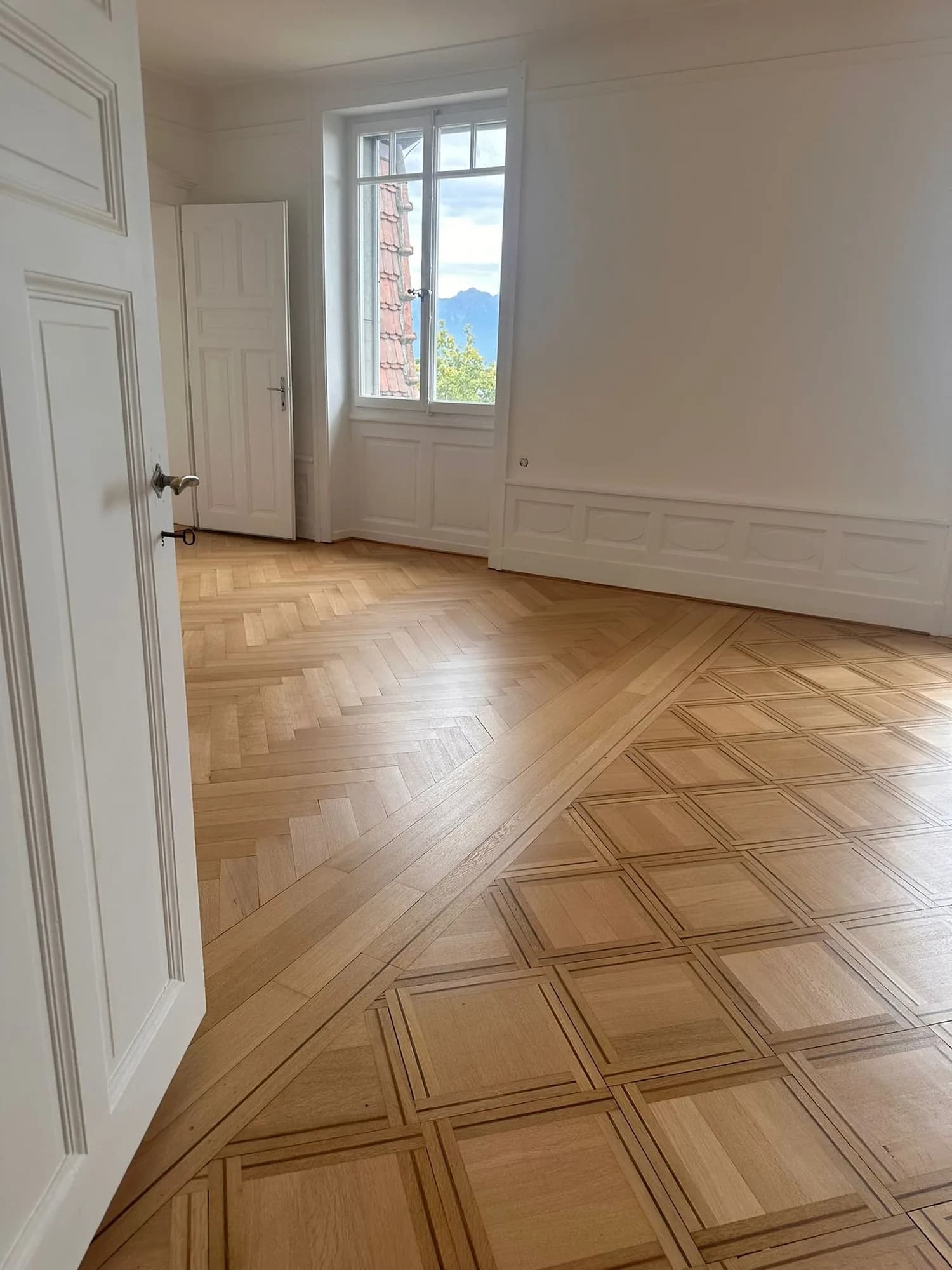 Pièce aux boiseries blanches : parquet en chevrons et parquet à panneaux séparés par une frise, fenêtre sur les montagnes