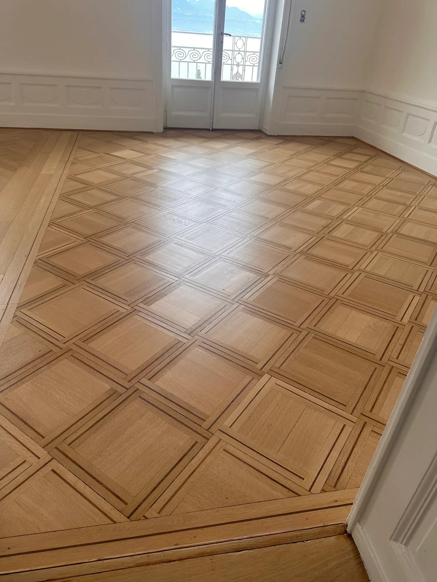Parquet à panneaux en bois clair dans une pièce aux murs blancs, porte-fenêtre au fond